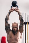 Poderoso fisiculturista sem camisa com tatuagens fazendo exercício com kettlebell pesado durante o treinamento funcional em ginásio — Fotografia de Stock