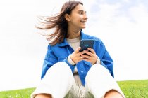 Baixo ângulo de feliz fêmea sentada na colina na natureza no dia ventoso e navegar na Internet no telefone celular enquanto olha para longe — Fotografia de Stock