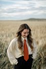Positive junge Frau in weißem Hemd und roter Krawatte, die Hände hinter dem Rücken zwischen Weizenspitzen auf dem Land stehend — Stockfoto