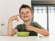Портрет очаровательного веселого мальчика, сидящего за обеденным столом дома и делающего жесты с широкой улыбкой — стоковое фото