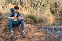 Seitenansicht von zufriedenen männlichen Wanderer mit Rucksack sitzt in der Nähe des Sees im Wald und surft Handy während der Reise — Stockfoto