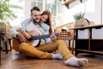 Веселый татуированный мужчина-музыкант, играющий на гитаре рядом с содержанием любимой женщины, глядя друг на друга в кресле в гостиной — стоковое фото