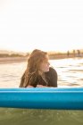 Женщина-серфер лежит на доске SUP и плавает на спокойной воде моря в солнечный день, глядя в сторону во время заката — стоковое фото