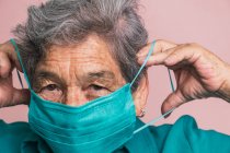Sorrindo a boca de cobertura feminina envelhecida com máscara médica de proteção azul do coronavírus enquanto olha para a câmera no fundo rosa no estúdio — Fotografia de Stock
