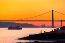 Argine affollato situato vicino al fiume Tago con traghetto vicino silhouette di 25 de Abril Ponte contro cielo tramonto arancione a Lisbona, Portogallo — Foto stock
