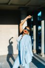 Вид сбоку возбужденной этнической женщины в стильной одежде и солнцезащитных очках, развлекающейся на городской тротуаре под солнечным светом — стоковое фото
