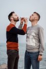 Homosexual male partners with modern haircuts enjoying champagne from glasses while standing on ocean coast in daytime — Stock Photo
