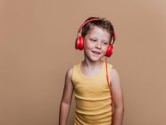 Satisfeito menino pré-adolescente em fones de ouvido vermelhos ouvindo música em fundo marrom em estúdio — Fotografia de Stock