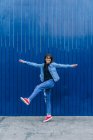 Hipster féminine excitée en tenue denim sautant avec les bras tendus sur fond bleu en ville et regardant la caméra — Photo de stock