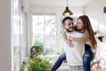 Smiling woman riding piggyback on boyfriend while looking at each other against windows at home — Stock Photo