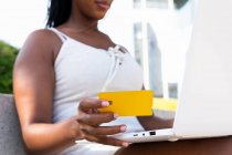 Niedriger Winkel einer anonymen Afroamerikanerin, die mit Plastikkarte bezahlt, während sie ihren Laptop beim Online-Shopping in der Straße von Barcelona benutzt — Stockfoto