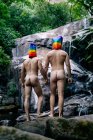 Vue de dos des hommes gays nus anonymes avec des sacs arc-en-ciel sur les têtes tenant la main tout en se tenant près de la chute d'eau dans la forêt — Photo de stock