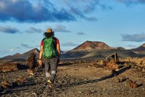 Randonneur anonyme avec sac à dos marchant sur un sentier sec dans la vallée près des collines par une journée nuageuse d'été à Fuerteventura, Espagne — Photo de stock