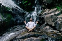 Homme paisible assis à Padmasana avec les mains mudra et les yeux fermés tout en faisant du yoga et en méditant sur la roche humide près de la cascade — Photo de stock