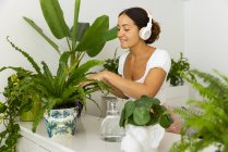 Gentle ethnic female listening to music from wireless headphones while touching tropical plant foliage in ornamental pot at home — Stock Photo