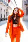 Stilvolle Frau mit Ingwerhaaren und in leuchtend orangefarbenem Anzug, die durch die Straßen der Stadt läuft und in die Kamera blickt — Stockfoto
