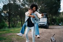 Smiling black woman riding piggyback on boyfriend while taking self portrait on cellphone against dog and caravan in camp — Stock Photo