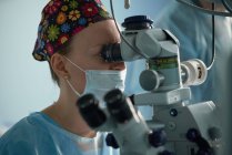 Medico donna adulto concentrato in maschera sterile e cappuccio medico ornamentale guardando attraverso il microscopio chirurgico contro il collega di coltura in ospedale — Foto stock