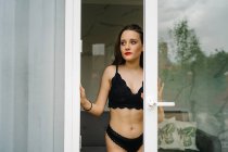 Conteúdo Feminino magro em lingerie preta em pé perto da porta de vidro que leva à varanda e olhando para longe — Fotografia de Stock