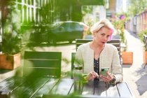 Альтернативна жінка з коротким волоссям, що переглядає соціальні медіа на смартфоні, сидячи за столом у вуличному кафе в сонячний день — стокове фото