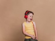 Soddisfatto ragazzo preadolescente in cuffie rosse ascoltare musica e firmare la canzone con gli occhi chiusi su sfondo marrone in studio — Foto stock
