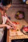 Vue de dessus de la femelle ethnique dans un tablier découpant des tomates mûres sur une planche à découper pendant la cuisson du déjeuner dans la cuisine à la maison — Photo de stock