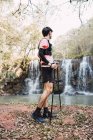 Вид збоку чоловічого мандрівника з палицями, що стоять біля озера з водоспадом у лісі — стокове фото