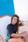 Femmina surfista seduta con bordo SUP blu sulla spiaggia sabbiosa in estate e guardando altrove — Foto stock