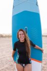 Surfista feminina feliz de pé com placa SUP azul na praia de areia no verão e olhando para a câmera — Fotografia de Stock