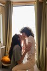 Вид сбоку на содержание ЛГБТ пара женщин целуются на диване дома и смотрят друг на друга с любовью — стоковое фото