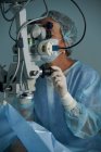 Взрослая женщина-врач в стерильной маске и декоративной медицинской шапке смотрит через хирургический микроскоп на коллегу по урожаю в больнице — стоковое фото