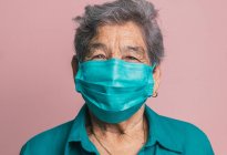 Усміхнена літня жінка використовує синю захисну медичну маску від коронавірусу, дивлячись на камеру на рожевому фоні в студії — стокове фото