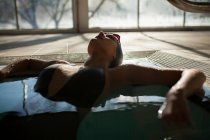 Jeune belle femme allongée sur le bord de la piscine intérieure, portant un maillot de bain noir — Photo de stock