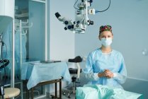 Взрослая женщина-врач в хирургической форме и стерильной маске смотрит в камеру, сидя в клинике — стоковое фото