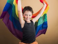 Fröhliches Kind mit geschminkten Wangen, das die LGBTQ-Flagge hisst, während es in die Kamera auf beigem Hintergrund blickt — Stockfoto