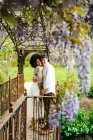 Vista lateral de una suave pareja multirracial abrazándose mientras está de pie con los ojos cerrados bajo el arco con flores de glicina violeta florecientes en el jardín - foto de stock