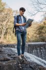 Bajo ángulo de excursionista masculino con mochila navegando con mapa de papel mientras está de pie en el bosque y mirando hacia otro lado - foto de stock