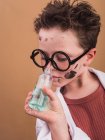Хімічна дитина в пластикових окулярах з плямами фарби на обличчі, що пахнуть рідиною з пляшки на бежевому фоні — стокове фото
