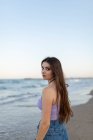 Dreamy young female with long hair looking at camera while standing on sandy beach near waving sea — Stock Photo