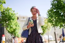 Веселая альтернативная женщина с окрашенными волосами, стоящая на улице и серфинг Интернет на мобильном телефоне летом — стоковое фото