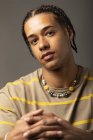 Молодий афроамериканець з плетеним волоссям, одягнений у збільшену смугасту сорочку та намисто, дивиться на камеру на сірий фон. — стокове фото