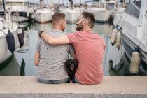 Rückenansicht des Hundes zwischen einem fröhlichen bärtigen Mann, der einen anonymen homosexuellen Partner umarmt, während er redet und auf einem Steg im Hafen sitzt — Stockfoto