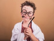 Bambino con macchie di vernice sul viso e occhiali tempio in accappatoio di laboratorio guardando la fotocamera su sfondo beige — Foto stock