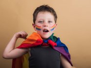 Fröhliches Kind mit geschminkten Wangen und LGBTQ-Flagge vor der Kamera auf beigem Hintergrund — Stockfoto