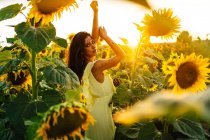 Anmutige junge hispanische Frau in stylischem gelben Kleid steht mit erhobenen Armen inmitten blühender Sonnenblumen auf einem Feld in einem sonnigen Sommertag und blickt in die Kamera — Stockfoto