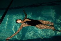 Giovane bella donna all'interno della piscina coperta, indossa un costume da bagno nero, galleggiante sulla schiena — Foto stock
