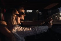 Vista lateral do casal amoroso em óculos de sol abraçando no carro vintage enquanto cavalga na natureza à noite — Fotografia de Stock