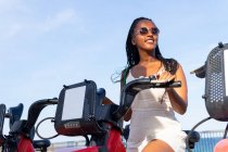 Vista lateral da mulher afro-americana elegante em pé perto de bicicleta alugada no passeio marítimo em Barcelona no verão — Fotografia de Stock