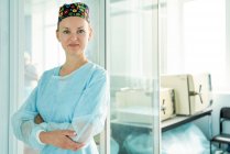 Самоуверенная взрослая женщина-врач со сложенными руками в декоративной медицинской шапке, смотрящая на камеру напротив стеклянной стены в больнице — стоковое фото