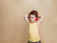 Niño preadolescente despreocupado en auriculares rojos escuchando música mientras está de pie sobre un fondo marrón y mirando a la cámara - foto de stock
